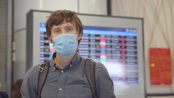 A Young Man Wearing a Medical Face Mask in an Airport Stands in a Hall in Front of a Flight Schedule