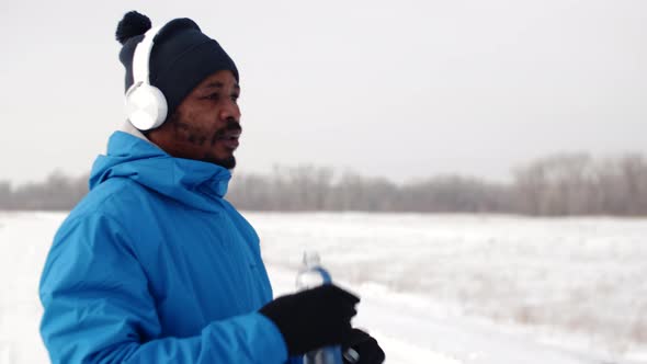 Black Man Wearing Headphones and Winter Sportswear Stops After Jogging and Drinks Water From Bottle