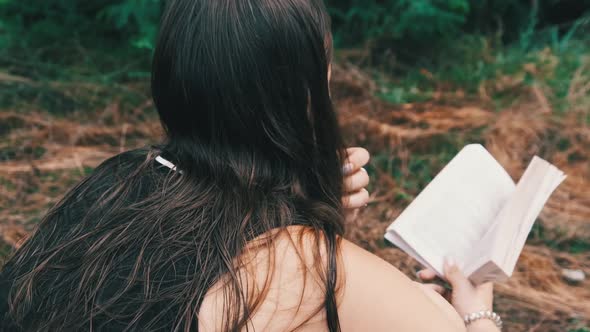 Young Woman Reads a Book Sitting on the Grass in Nature at Summer Day