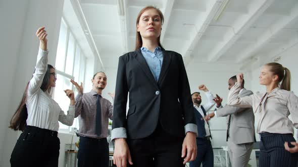 Low Angle Shot of Ambitious Woman Standing in Office with Serious Face While Coworkers Dancing