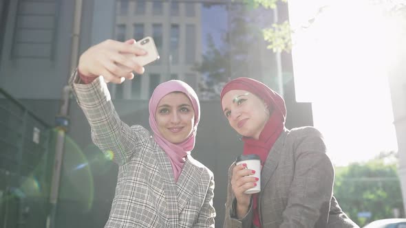 Two Young Muslim Women Wearing Hijab Headscarf Making Selfie at Phone, Sitting Together at Bench