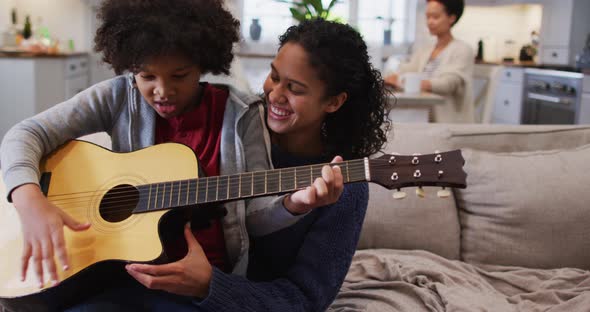 Mixed race mother and daughter sitting on couch playing guitar