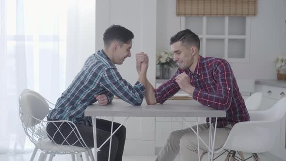 Caucasian Twin Brothers Competing in Armwrestling at Home. Two Positive Adult Brunette Men Sitting