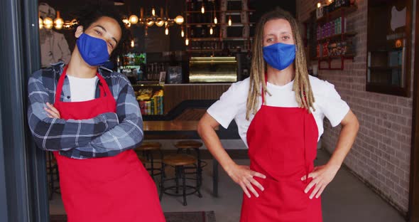 Two diverse male baristas wearing face masks and aprons standing in doorway of cafe