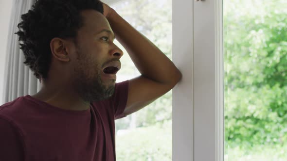 Profile of tired biracial man standing at window and yawning