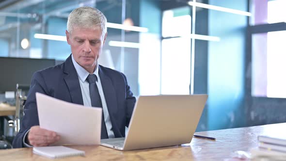 Middle Aged Businessman Reading Document and Using Laptop