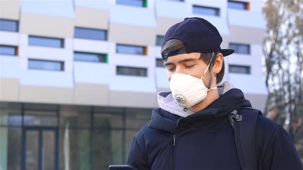 Young Man in Protective Respiratory Mask Using Smartphone on the Street in City