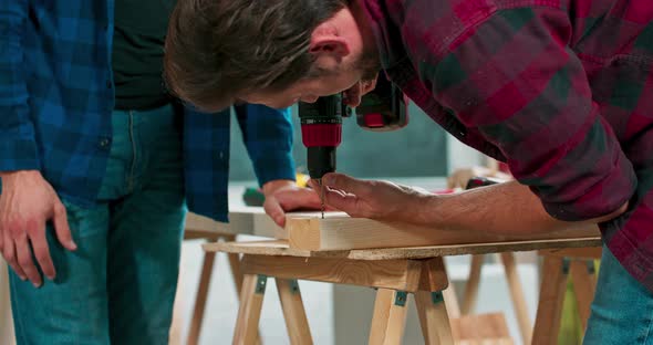 Concentrated Young Bearded Carpenter Wearing Jeans and a Plaid Flannel Shirt Drilling a Hole in a