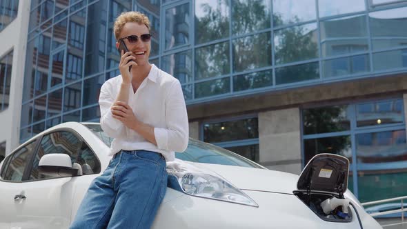 Stylish Modern Young Curly Man Charges His Electric Car in the Yard of a Residential Complex and