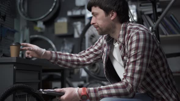 A Tired Bicycle Mechanic Drinks Coffee at His Workplace While Calculating the Cost of Working in the