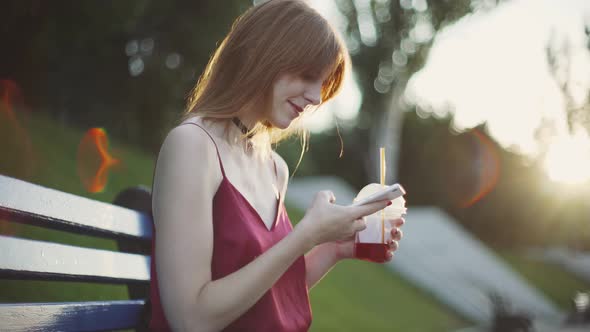 Beautiful Young Redhaired Woman Sitting on a Bench in the Park Drinking Juice and Using a Smartphone