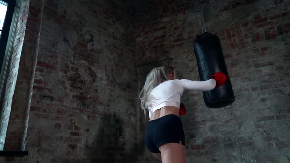 a Blonde with Long Hair in a White Top and Red Boxing Gloves Trains with a Punching Bag Against a