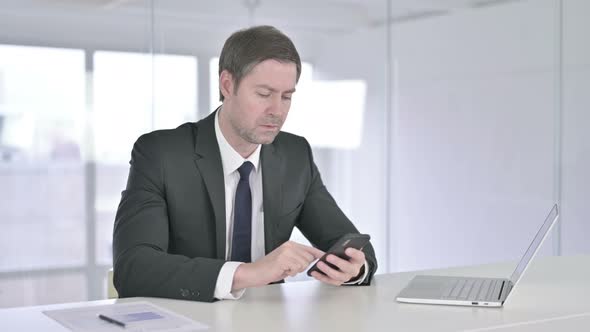 Cheerful Middle Aged Businessman Using Smartphone in Office