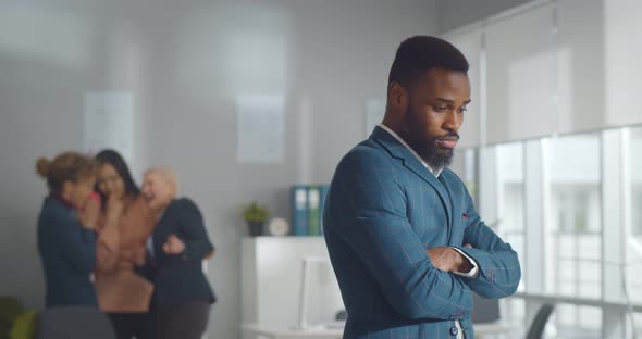 Portrait of Sad African Businessman Standing in Office with Female Colleagues Laughing at Him on