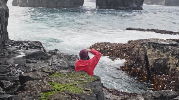 Man in Red Jacket Sitting on Rocks Taking a Picture of Sea Waves in Drangarnir