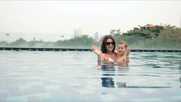 Portrait of Beautiful Mother with Her Son Standing in Swimming Pool and Laughing During Vacation on