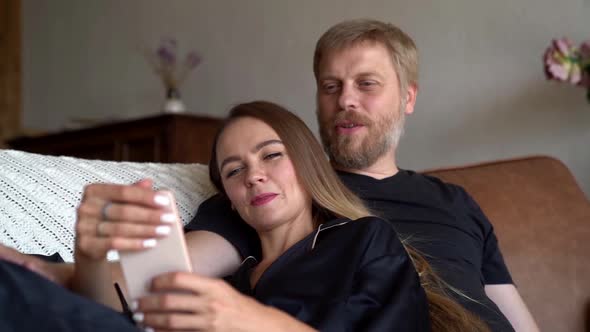 A Happy Married Couple Uses a Mobile Phone and Cuddles on the Sofa at Home. Husband and Wife Relax