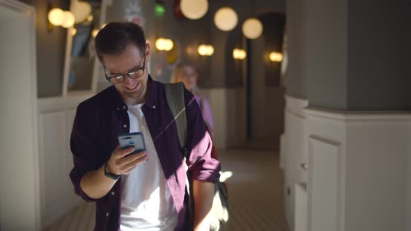 Cheerful Male Student Surfing Internet on Smartphone Walking in University Hall