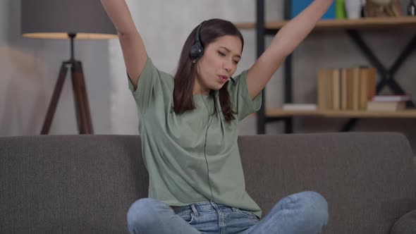 Joyful Excited Young Asian Woman in Earphones Rejoicing Startup Strategy Success Sitting on Couch in