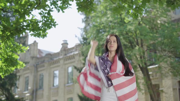 Beautiful Brunette Woman Wrapped in American Flag Raising Hand Up. Portrait of Confident Charming