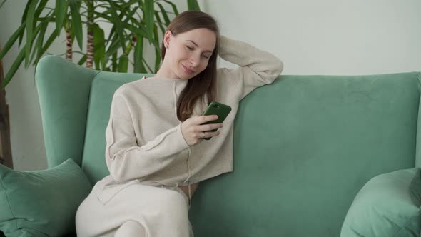 Smiling Woman Relaxes Sitting on the Couch at Home Using Mobile Phone Text Messages To Chat