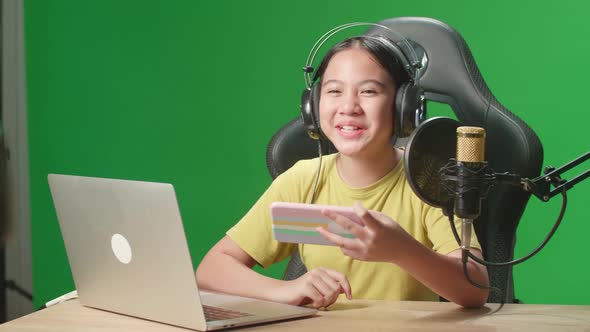 Asian Kid Girl Holding Mobile Phone And Talking To Camera While Live Stream On Green Screen