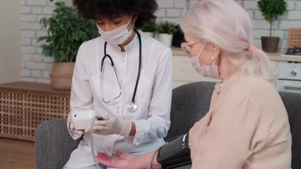 Afro American Woman Doctor in Mask Checks Blood Pressure Senier Woman at Home Sitting on Couch