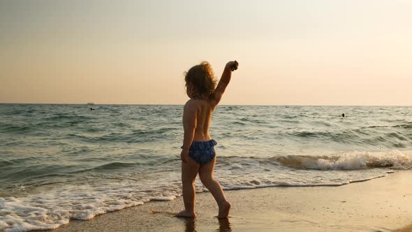 Little Funny Blonde Girl with Curly Hair in Swimming Suit Comes Up to Sea or Ocean at Sunset and