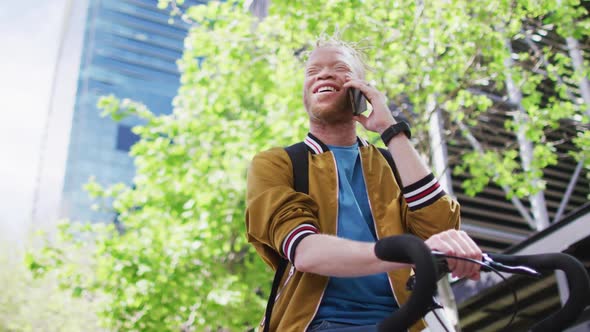 Happy albino african american man with dreadlocks in park with bike talking on smartphone