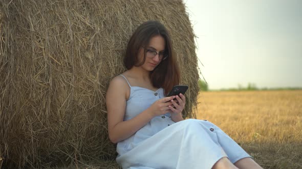 A Young Woman Sits Near a Haystack in a Field with a Phone in Her Hands