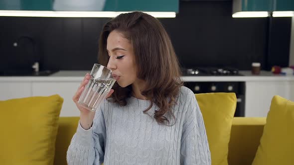 Happy Woman with Wavy Hair Sitting on the Couch in the Cuisine and Drinking a Glass of Pure Water