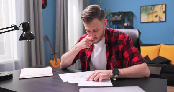 A Tired Young Man Sits at His Desk in His Room and Reviews His Apartment alt