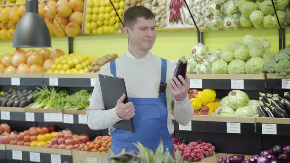 Young Positive Caucasian Employee Posing in Grocery with Folder and Eggplant. Portrait of Smiling