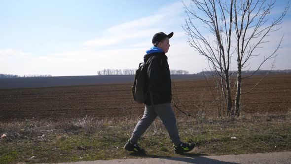 Happy Male Child with Backpack Goes Along a Driveway Near Ploughed Meadow. Small Kid in Cap Moves on