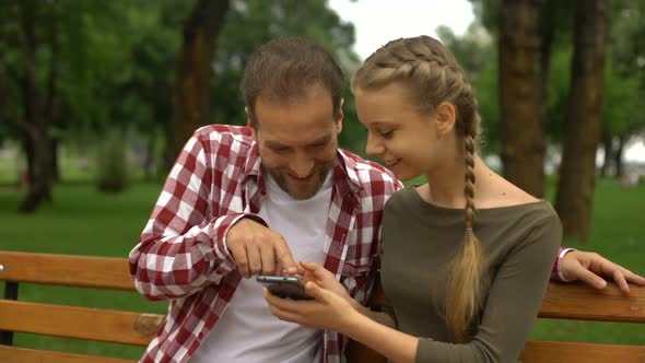 Cute Teenage Daughter Showing Funny Video on Cellphone to Father, Relax in Park