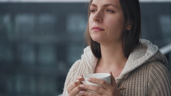 Caucasian Woman Stays on Balcony During Snowfall with Cup of Hot Coffee or Tea