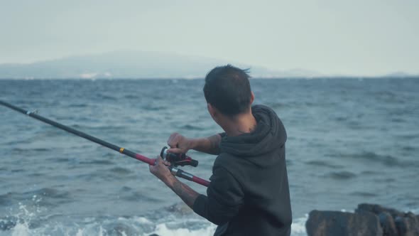 Young man struggling to real in fishing rod at windy seaside coastline