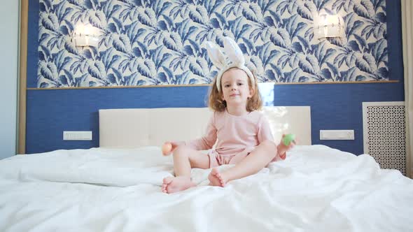 A Cute Little Girl with Long Hair Sits in Center of a Large Bed and Plays with Decorative Rabbit