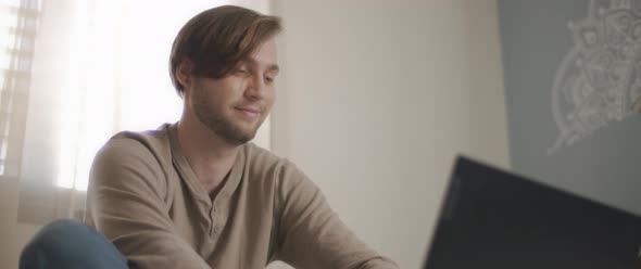 A close up of a young man smiling while sitting in bed and working on a computer. 