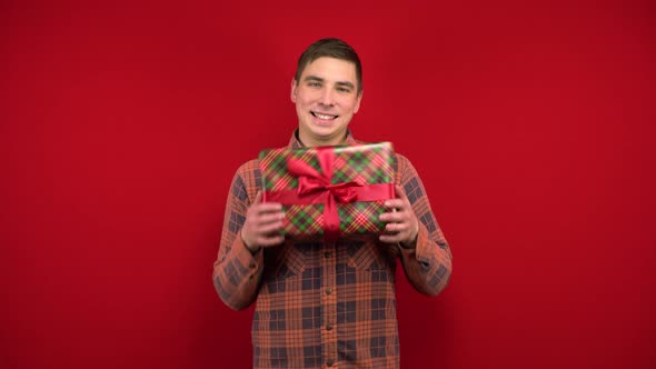 A Young Man Holds a Christmas Present and Rejoices. Shooting in the Studio on a Red Background.