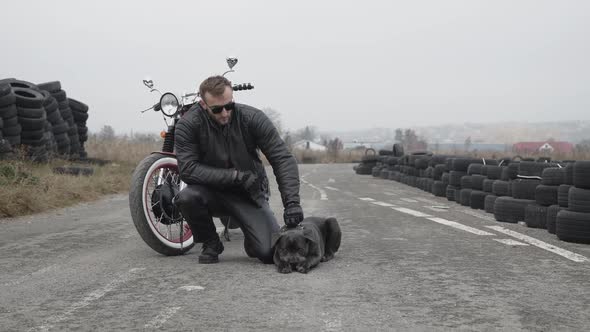 Potrait of Brutal Biker Holds Dark Bulldog at Motorcycle