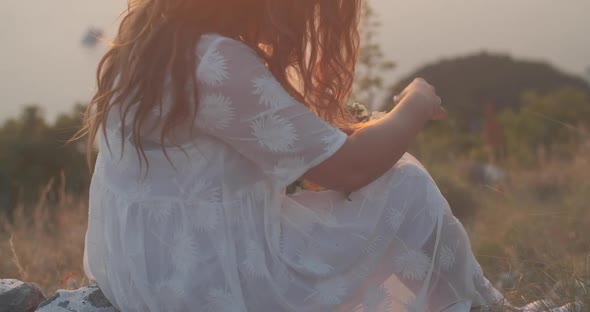 Woman in a white dress sits on a mountain and makes a wreath of flowers at sunset