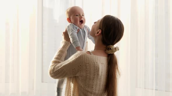 Happy Smiling Mother Lifting Up Playing and Kissing Her Baby Against Big Window at House