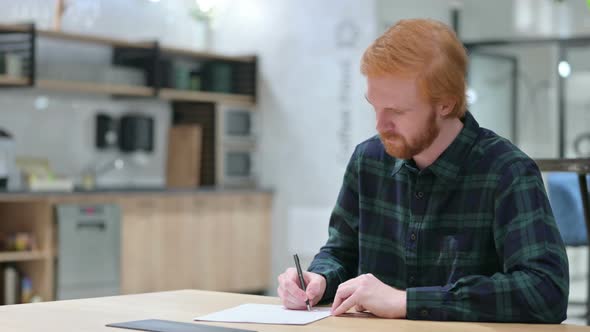 Young Beard Redhead Man Writing on Paper in Cafe 