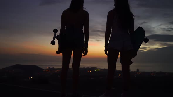 Two Girls Standing on the Edge of the Road with Skateboards in Their Hands Watching the Sunset