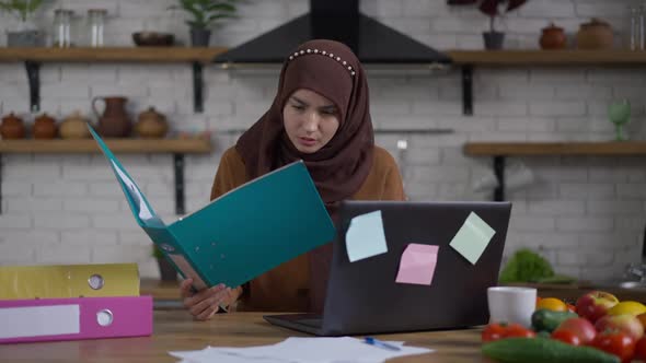 Focused Concentrated Woman in Hijab Comparing Documents and Online Information on Laptop