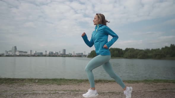 Slow Motion Young Sportswoman in Sportswear Trains in a Park Near the River