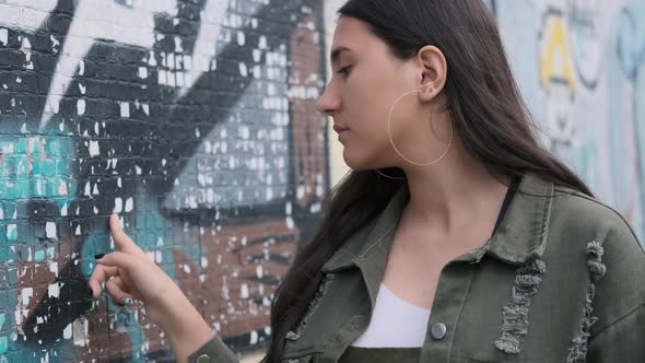 Thoughtful Young Brunette Girl Holds Her Hand on the Wall with Graffiti and Looks Into the Camera