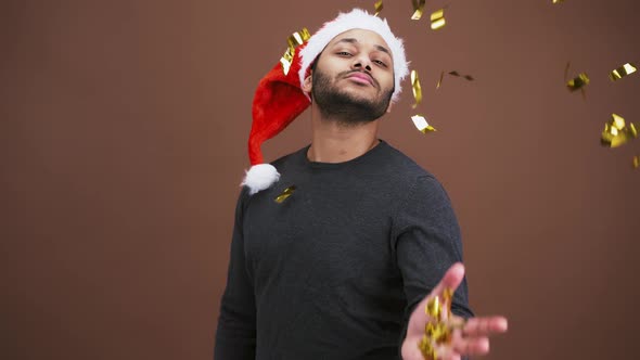 Arrogant Indian Man in Santa Hat Throwing Golden Glitter to Camera Brown Studio Background Slow