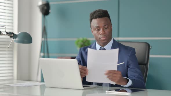 African Businessman with Laptop Reading Documents in Office